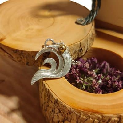 Colgante personalizado “Udane” de Emeki Joyas, pieza de oro con tres ondas de plata que simbolizan a padre, madre e hija, y una esfera dorada que representa el sol y el nacimiento; presentado en caja de madera artesanal decorada con flores secas