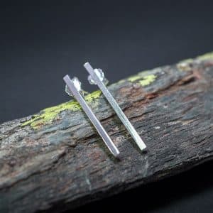 Pendientes de barritas minimalistas de Emeki Joyas en plata, realizados con hilo cuadrado y dispuestos en composición vertical, sobre piedra con líquenes