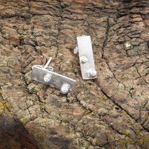 Pendientes Xirimiri Txiki de Emeki Joyas, piezas pequeñas de joyería artesanal en plata reciclada con geometría serena, apoyadas sobre tronco de madera.