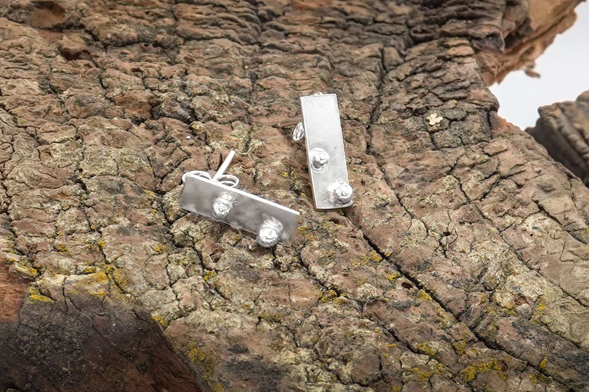 Pendientes Xirimiri Txiki de Emeki Joyas, piezas pequeñas de joyería artesanal en plata reciclada con geometría serena, apoyadas sobre tronco de madera.